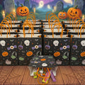 A Halloween-themed display of black treat bags with orange handles, decorated with skulls, pumpkins, ghosts, and candy motifs. A small matching tablecloth and orange bags are arranged on a wooden floor in front of a spooky graveyard scene with glowing pumpkins, tombstones, and floating ghosts under a purple night sky.