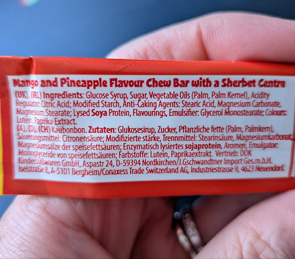 A close-up of a hand holding a red and orange package of Mango and Pineapple Flavour Chew Bar with a Sherbet Centre, showing the ingredients text in white on the side.