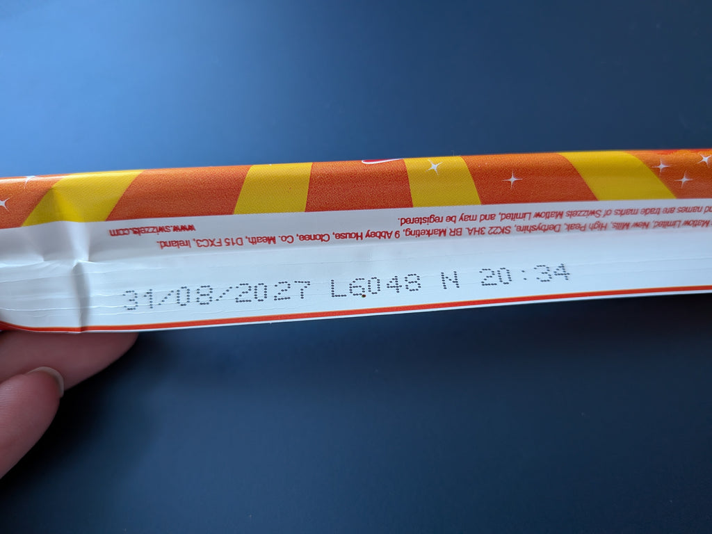 A close-up of a white paper strip with orange and yellow diagonal stripes, held by a hand against a dark blue background. The strip has printed text including the use by date.