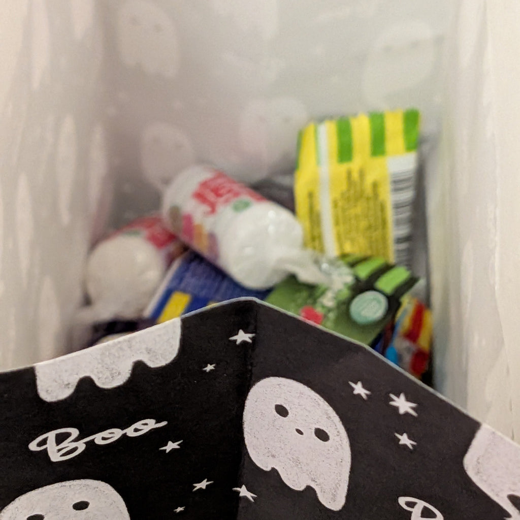 A close-up of a black Halloween-themed bag or container with white ghost and 'Boo' patterns, partially filled with assorted packaged snacks and drinks inside a white plastic bin.